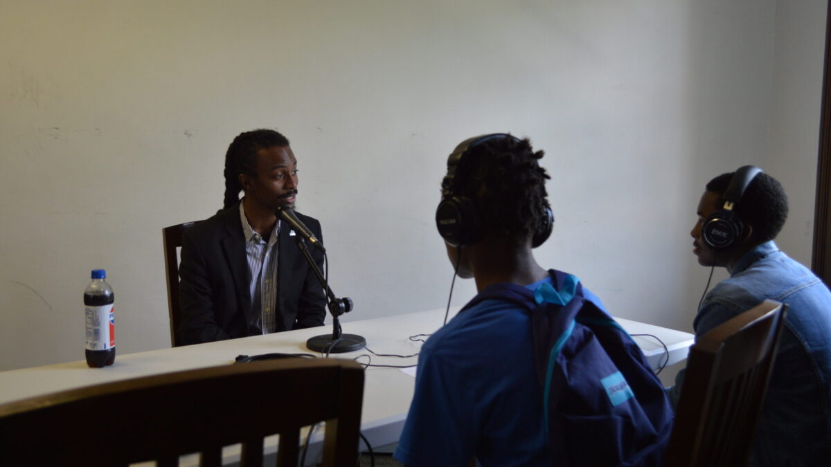 Two teens wearing headphones interviewing an adult speaking into a microphone on a white table