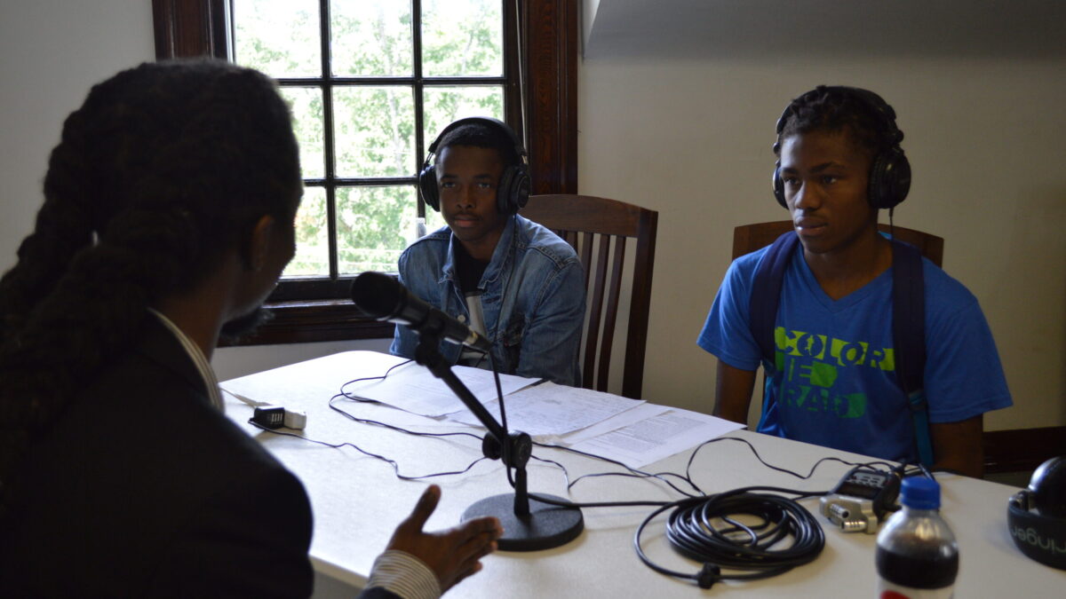 Two teens wearing headphones interviewing an adult speaking into a microphone connected to a portable recorder on a white table