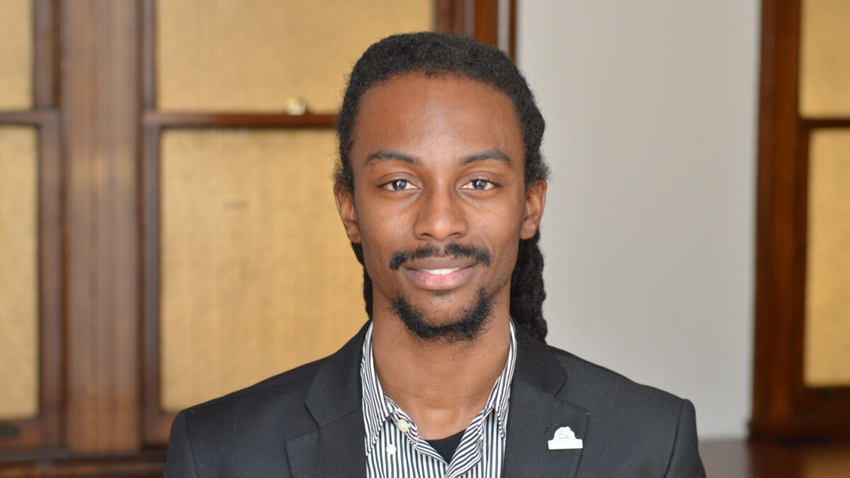 Adult with long black curly hair, a short mustache and beard, and a black suit jacket smiling in profile