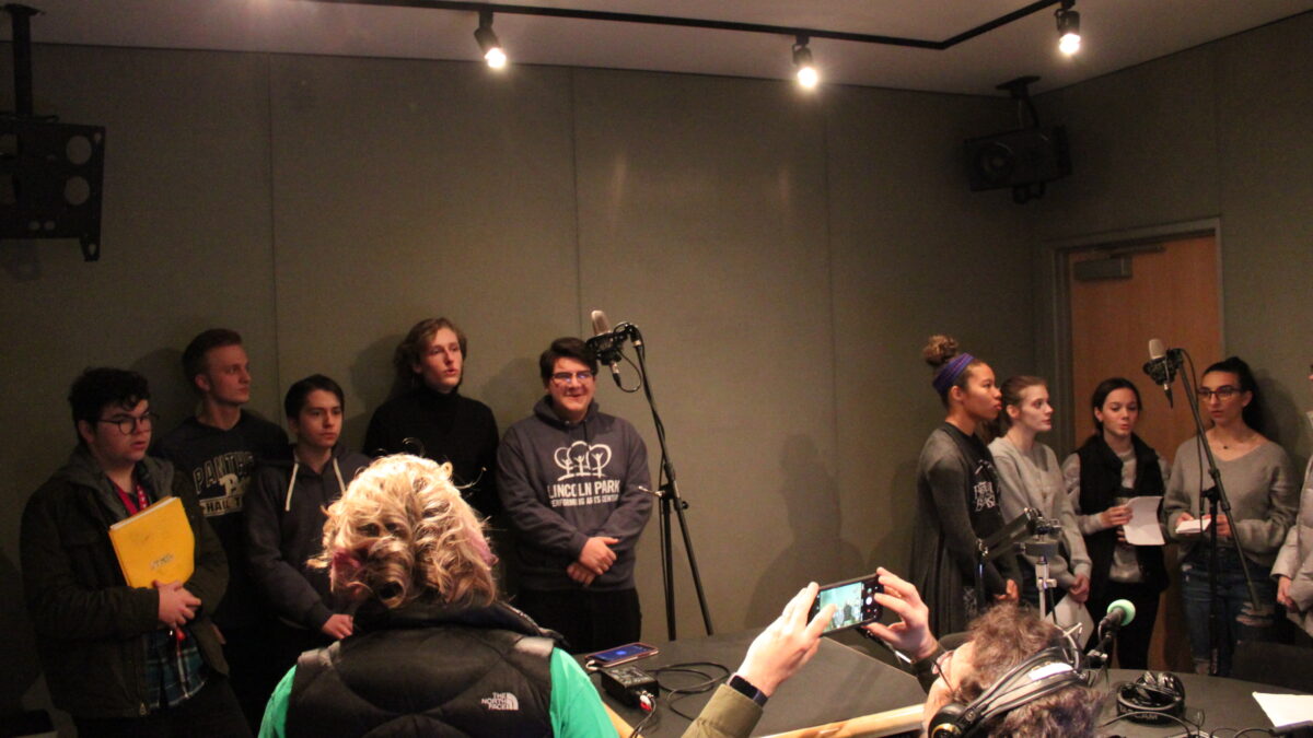 Ten teens singing around two microphones as an adult supervises and another adult wearing headphones takes a photo with their smartphone in a radio studio