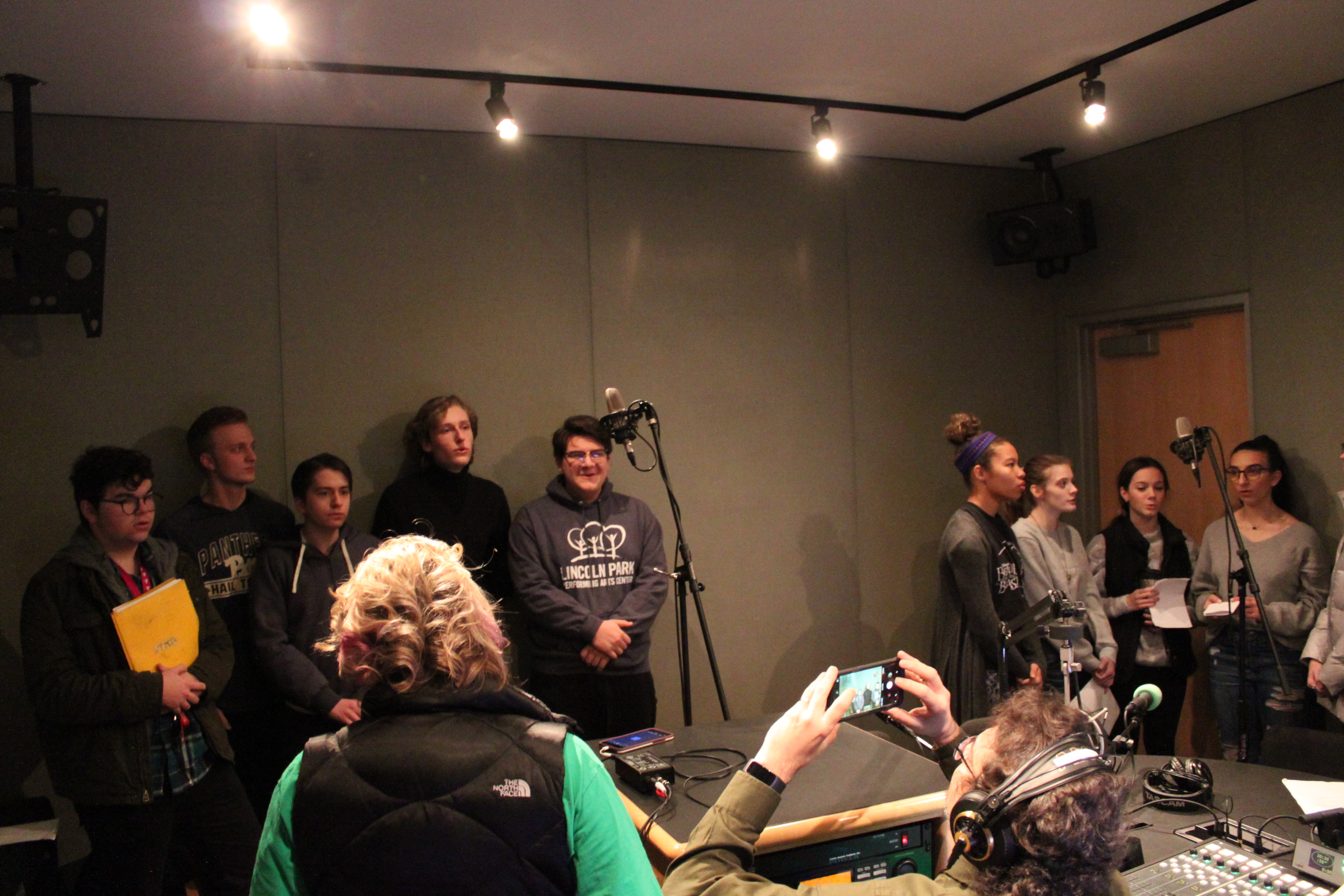 Ten teens singing around two microphones as an adult supervises and another adult wearing headphones takes a photo with their smartphone in a radio studio
