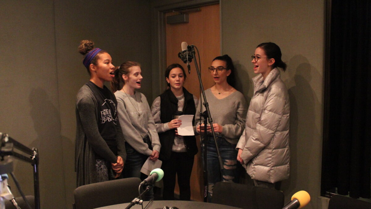 Five teens singing around a microphone in a radio studio