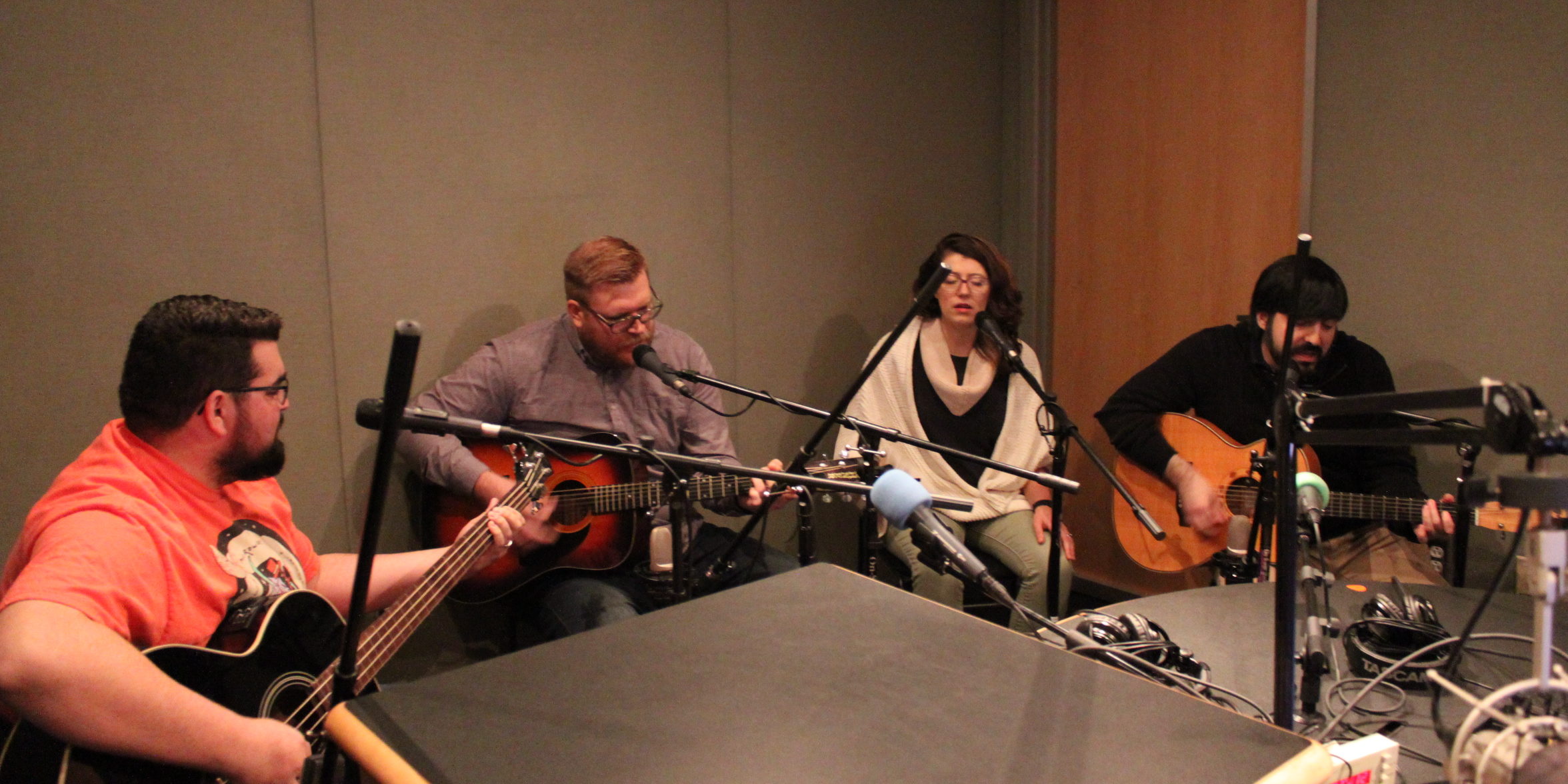 Four adults singing into microphones, three playing acoustic guitar, in a radio studio