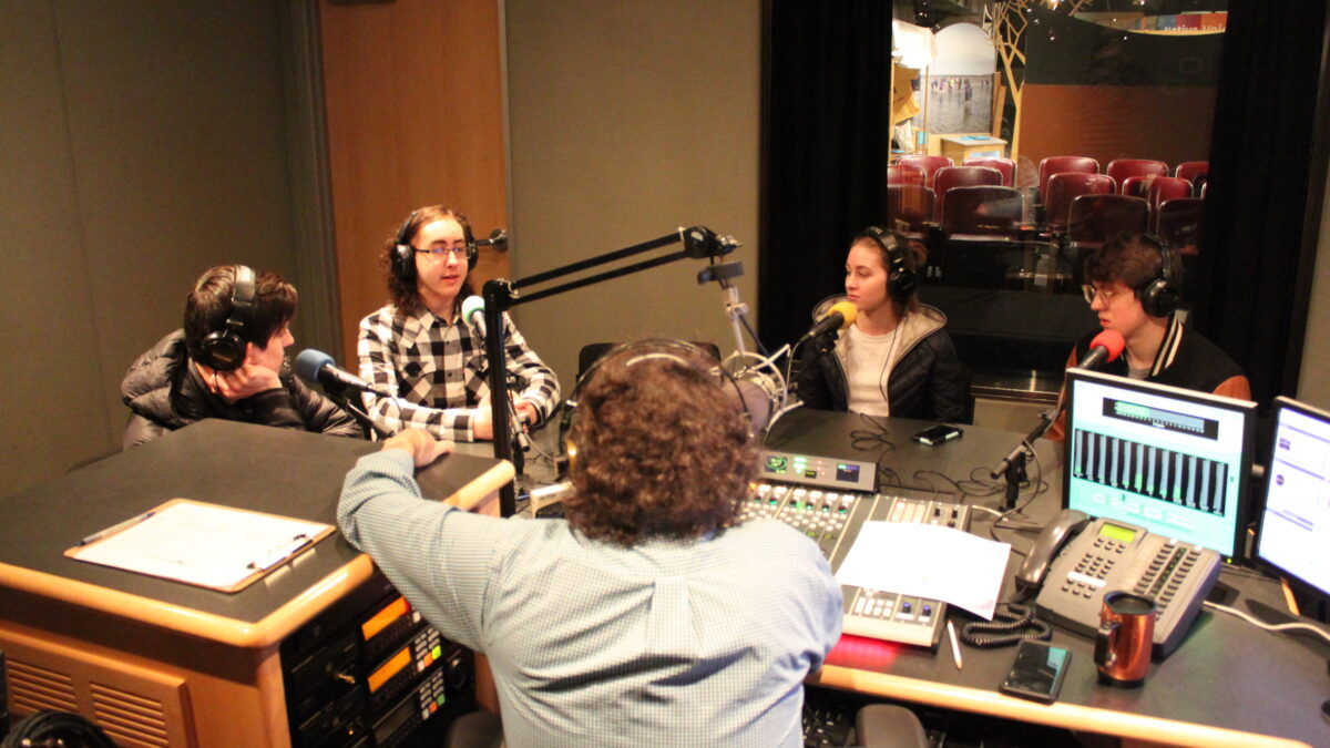 Four teens wearing headphones speaking into microphones being interviewed by an adult wearing headphones speaking into a microphone sitting behind a mixing board in a radio studio