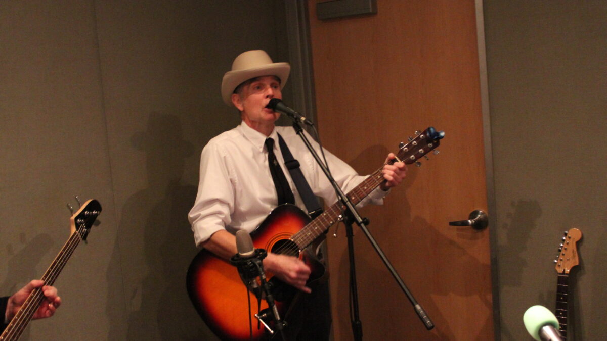 Adult playing acoustic guitar singing into a microphone in a radio studio