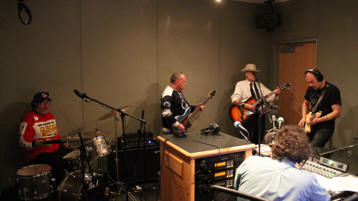 Adult wearing headphones playing drum set, adult playing bass, adult playing acoustic guitar, and an adult wearing headphones playing electric guitar, while another adult wearing headphones sitting behind a mixing board listens in a radio studio