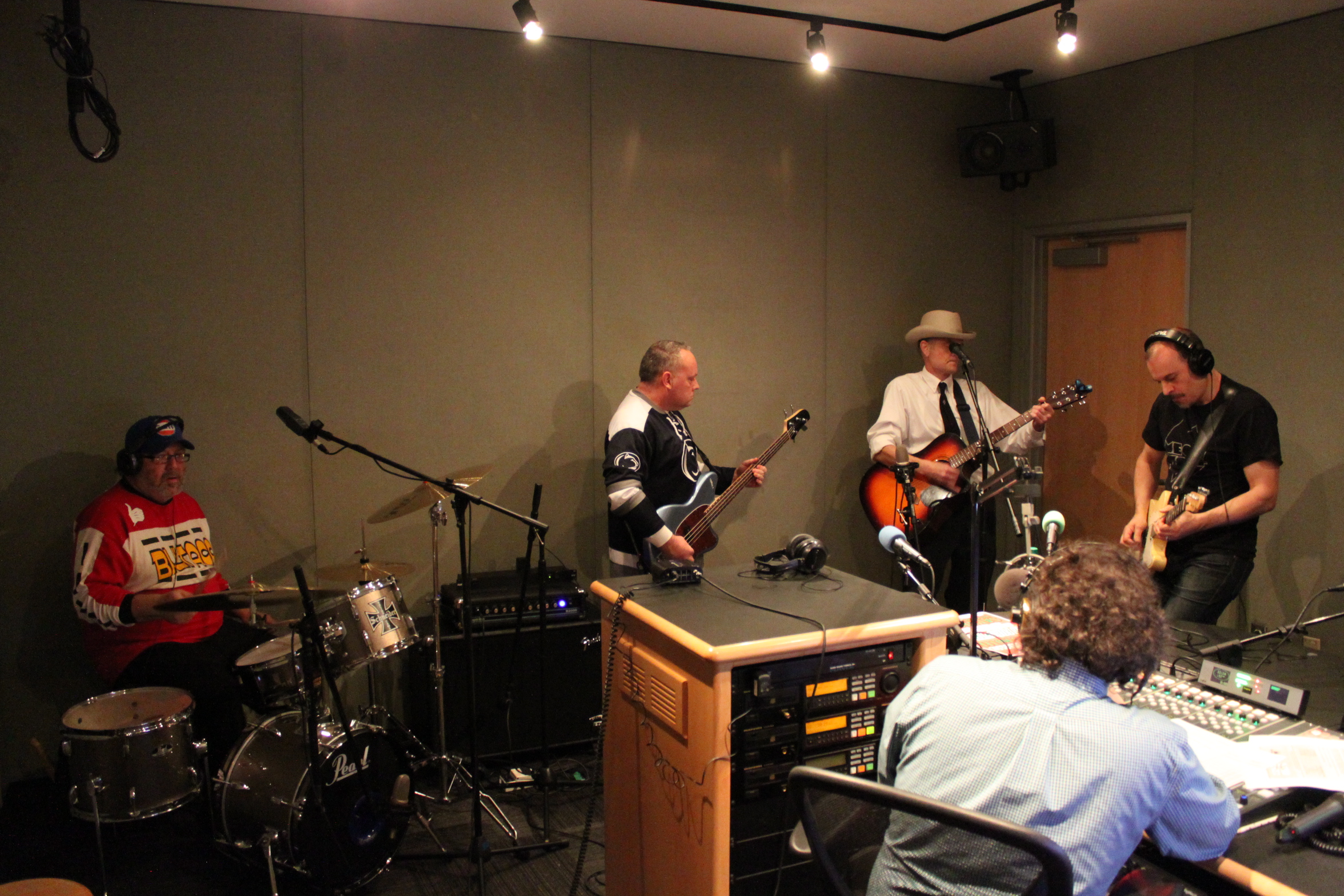 Adult wearing headphones playing drum set, adult playing bass, adult playing acoustic guitar, and an adult wearing headphones playing electric guitar, while another adult wearing headphones sitting behind a mixing board listens in a radio studio