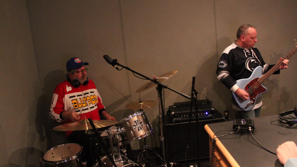 An adult wearing headphones playing drum set and an adult playing bass in a radio studio
