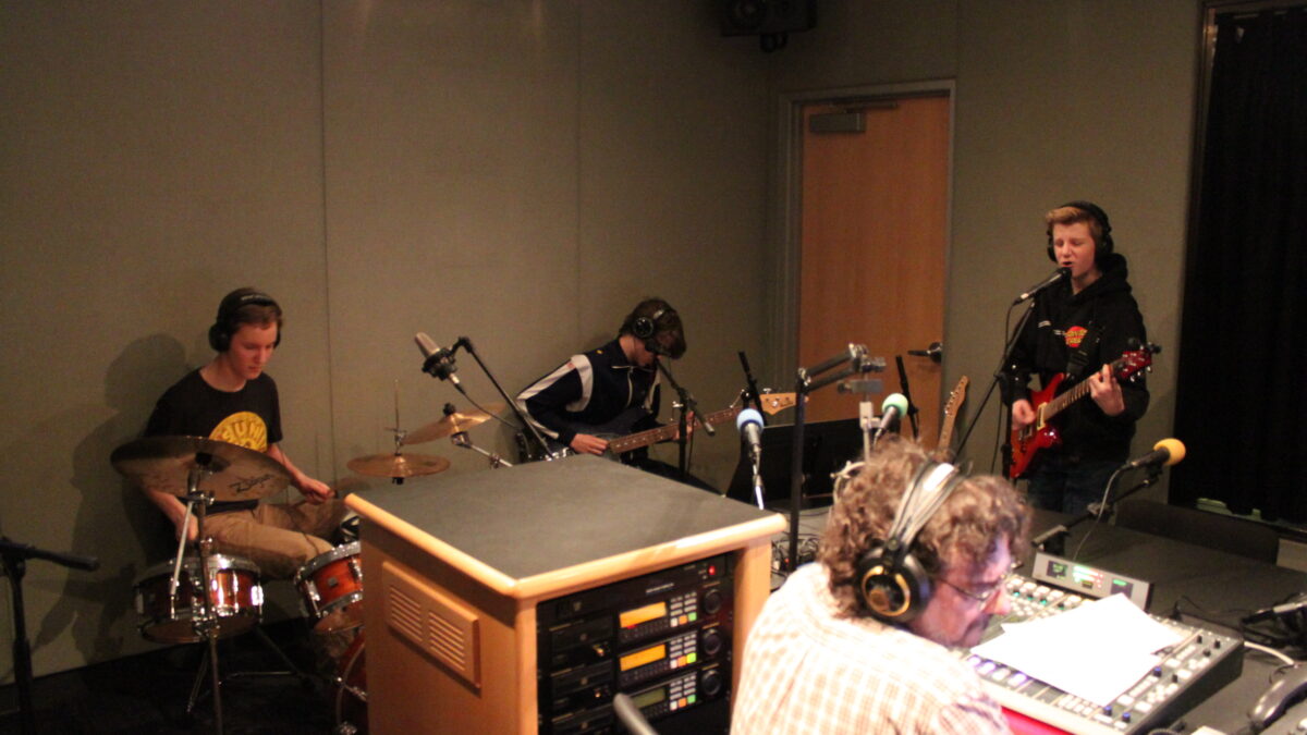 A teen wearing headphones playing drum set, a teen wearing headphones playing bass, a teen wearing headphones playing guitar and singing into a microphone, and an adult wearing headphones sitting behind a mixing board listening in a radio studio