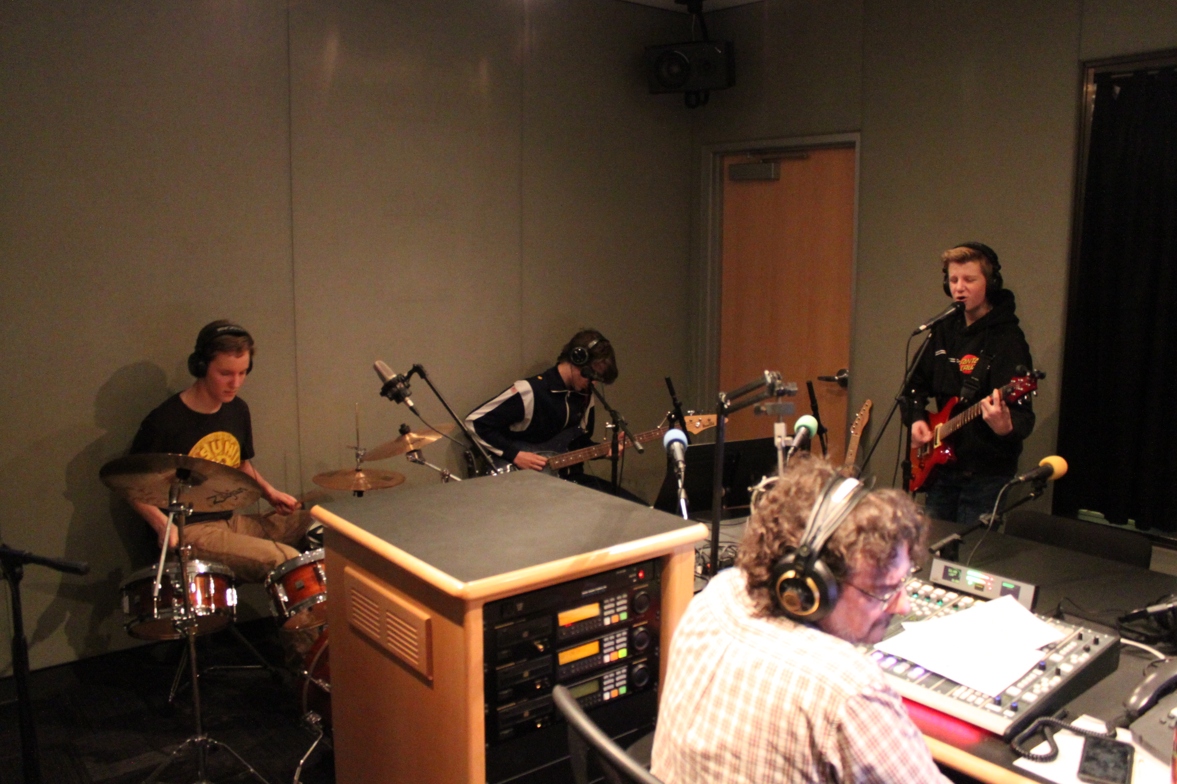 A teen wearing headphones playing drum set, a teen wearing headphones playing bass, a teen wearing headphones playing guitar and singing into a microphone, and an adult wearing headphones sitting behind a mixing board listening in a radio studio