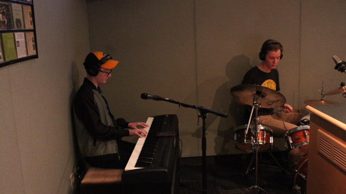 A teen wearing headphones playing piano keyboard and a teen wearing headphones playing drum set in a radio studio
