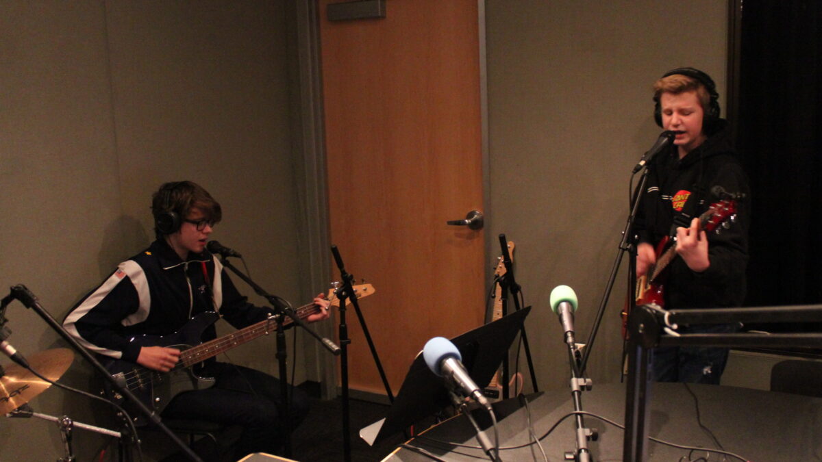 A teen wearing headphones playing bass and singing into a microphone and a teen wearing headphones playing guitar and singing into a microphone in a radio studio