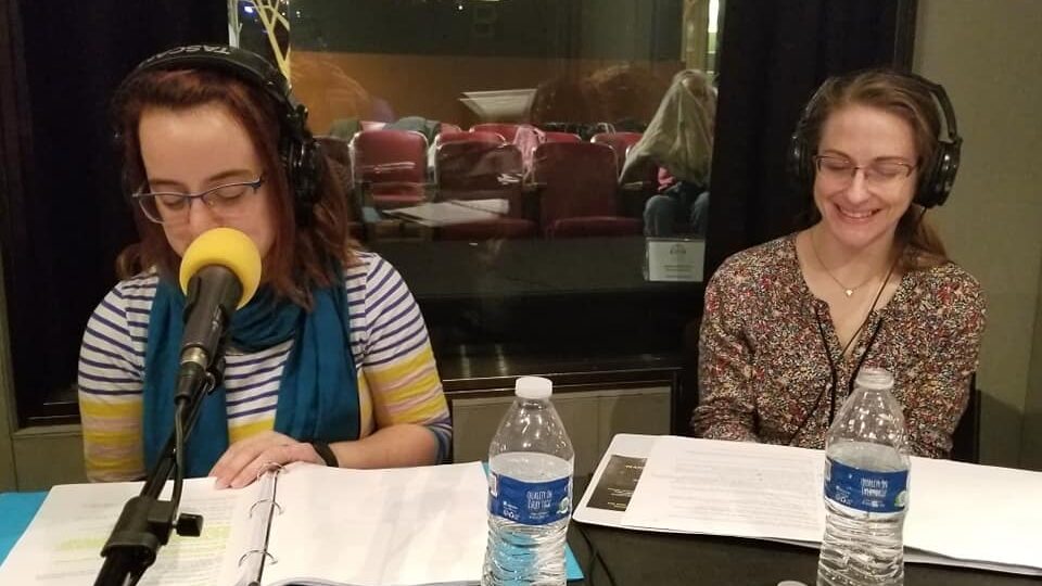 Two adults wearing headphones sitting in front of microphones and water bottles looking at paper in binders in a radio studio