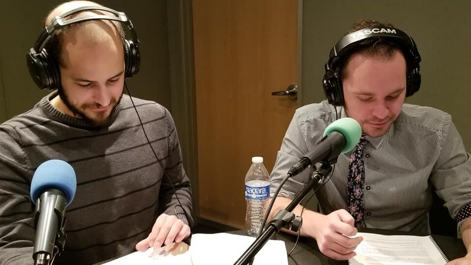 Two adults wearing headphones sitting in front of microphones looking down at pieces of paper in a radio studio