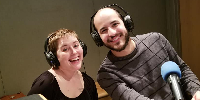 Two adults wearing headphones smiling in front of a microphone in a radio studio