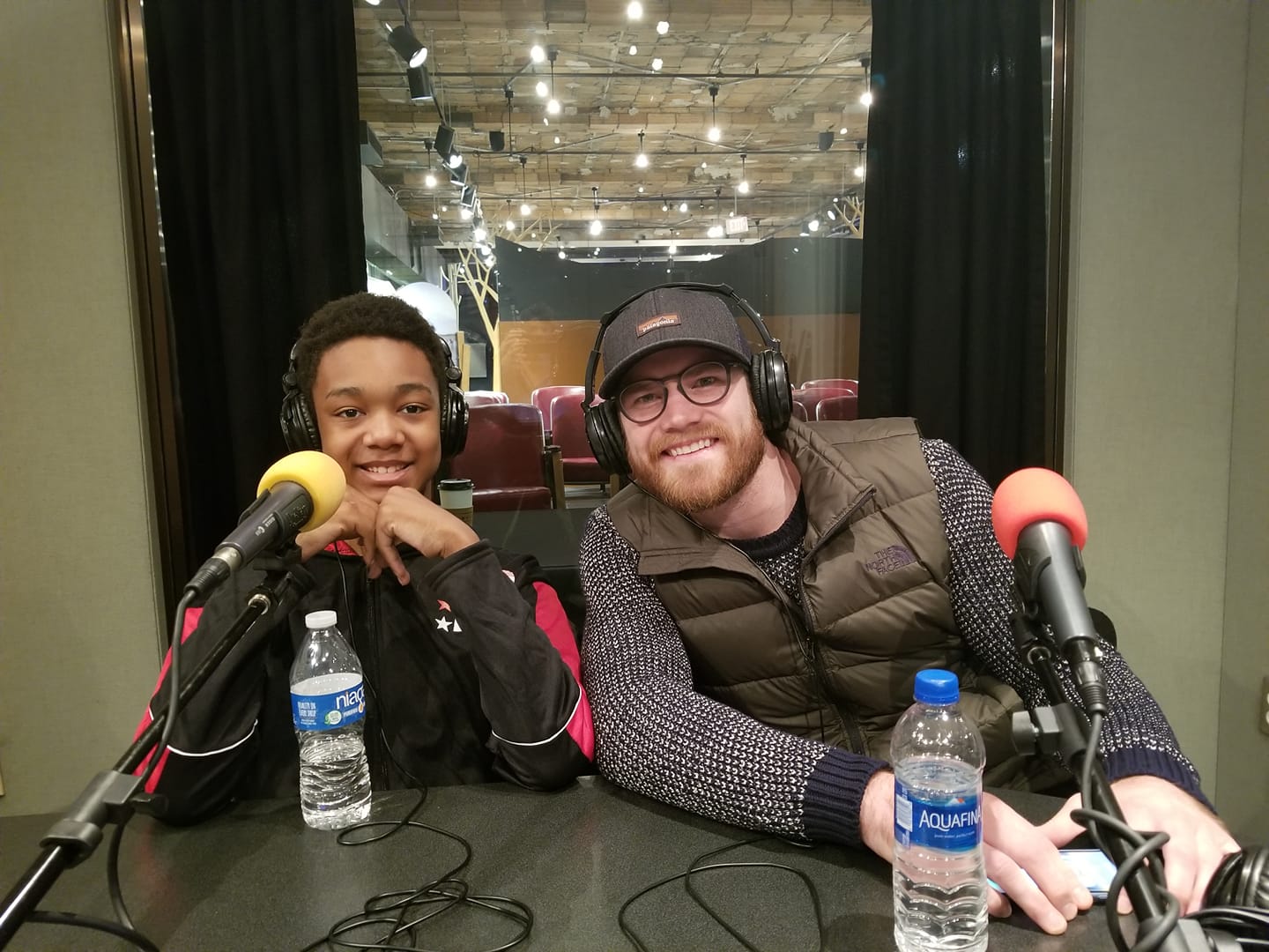 A youth and an adult both wearing headphones smiling behind microphones and bottles of water in a radio studio