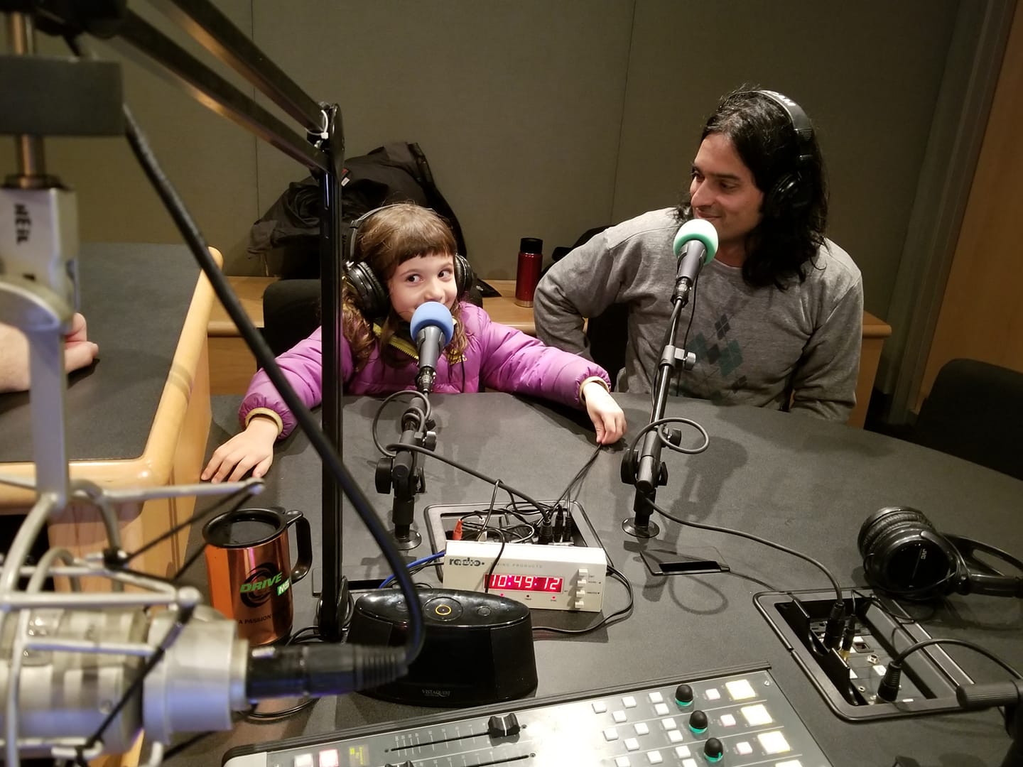A child and an adult wearing headphones sitting behind microphones in a radio studio