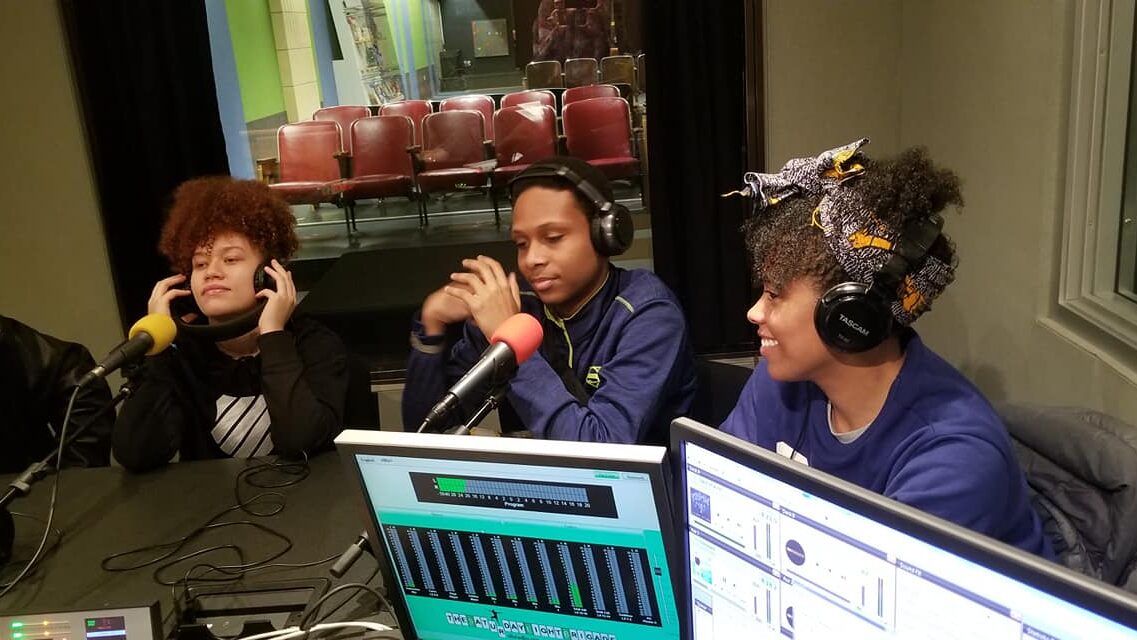 Three teens wearing headphones speaking into microphones in a radio studio