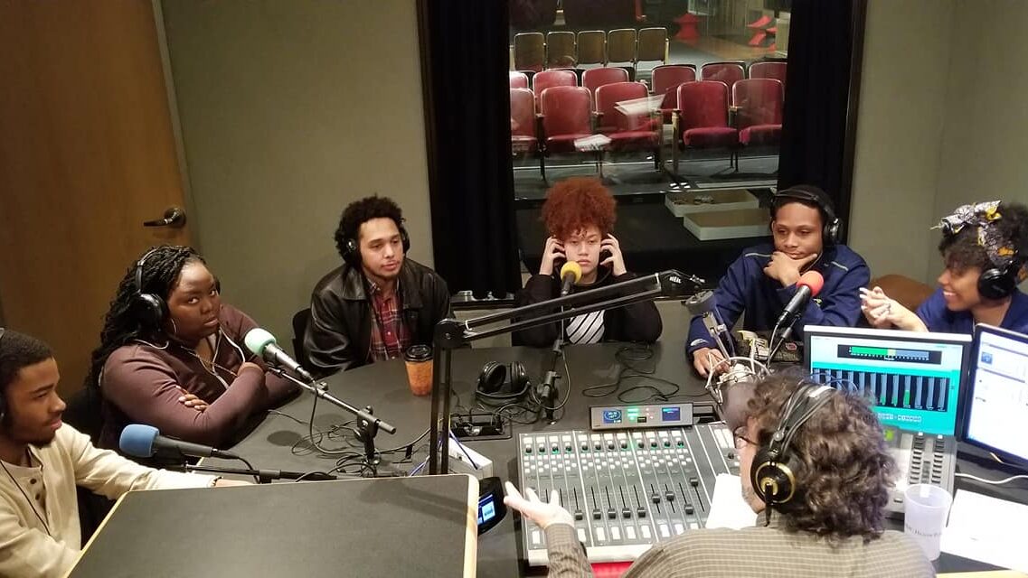 Six teens wearing headphones speaking into microphones being interviewed by an adult wearing headphones speaking into a microphone sitting behind a mixing board and gesturing with their hand in a radio studio