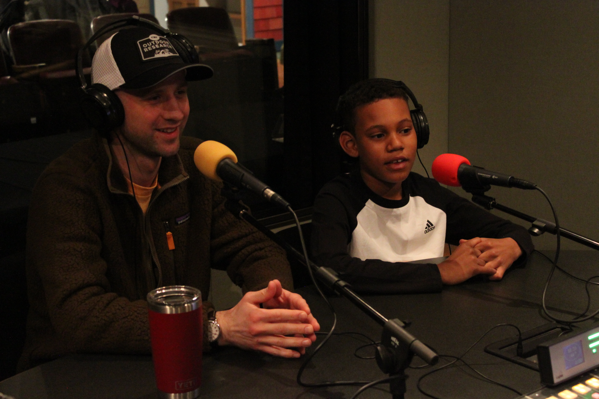 An adult and a child wearing headphones speaking into microphones in a radio studio