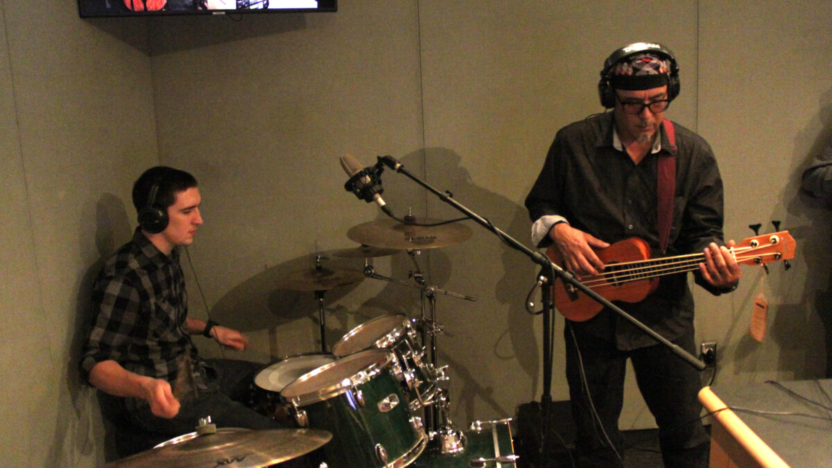 An adult wearing headphone playing drum set, and an adult wearing headphones playing bass ukulele in a radio studio
