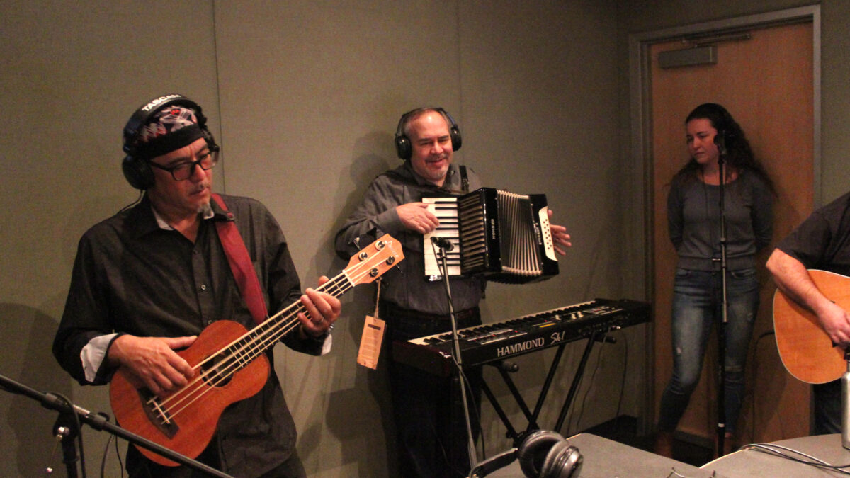 An adult wearing headphones playing bass ukulele, an adult wearing headphones paying accordion, and an adult standing behind a microphone in a radio studio