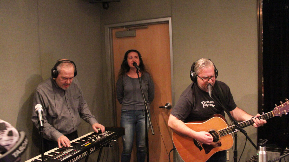 An adult wearing headphones playing synthesizer, an adult wearing headphones singing into a microphone, and an adult wearing headphones playing guitar and singing into a microphone in a radio studio
