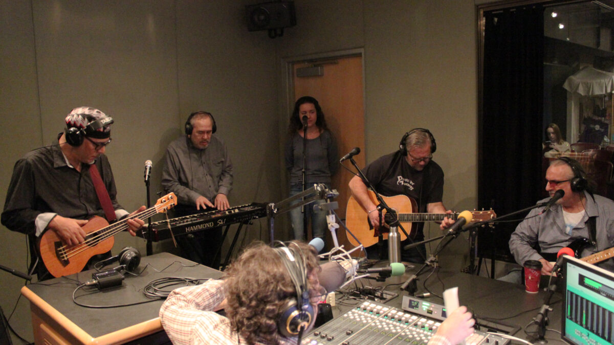 Five adults wearing headphones performing on bass ukulele, synthesizer, vocals, guitar, and bass, while another adult wearing headphones listens in a radio studio