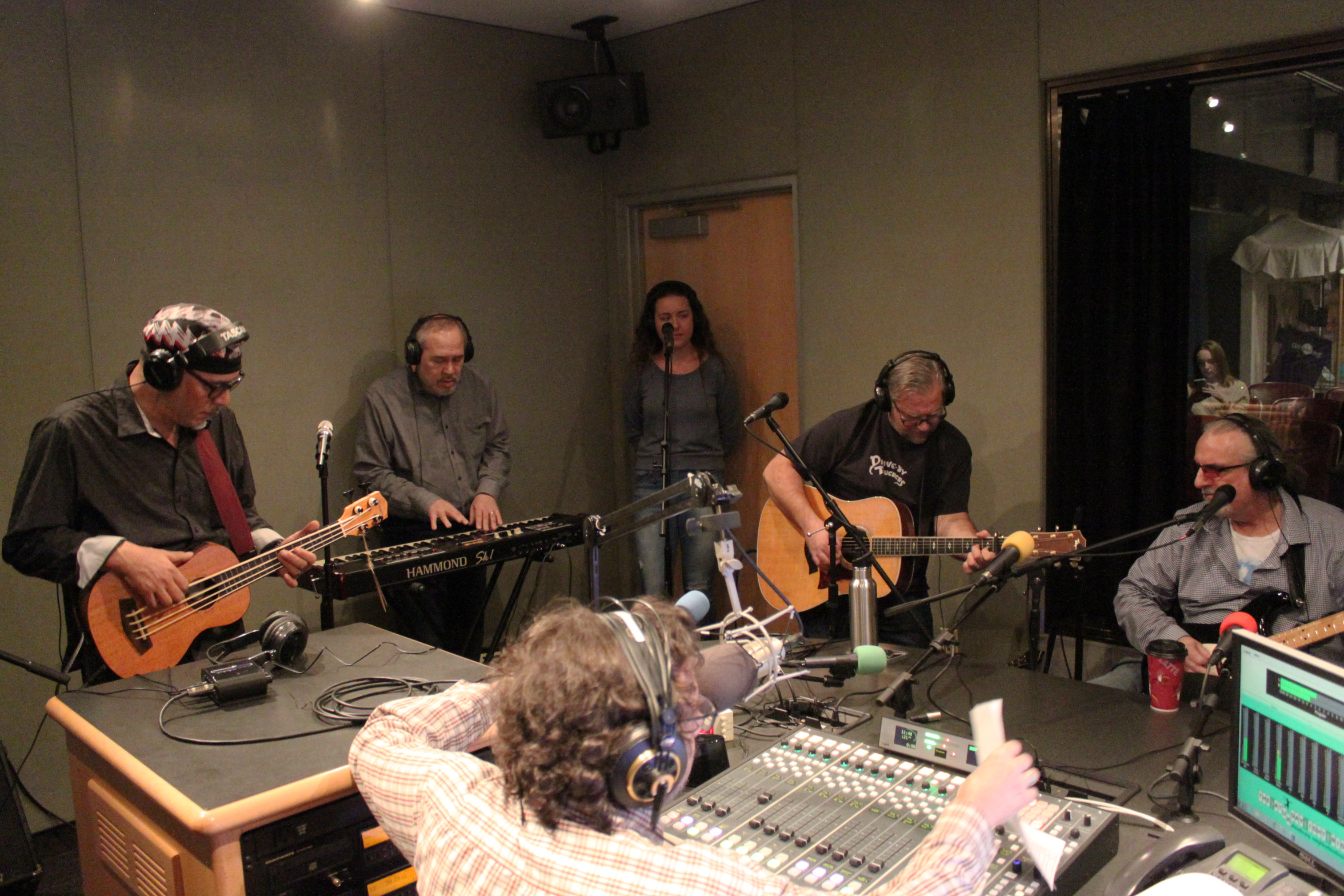 Five adults wearing headphones performing on bass ukulele, synthesizer, vocals, guitar, and bass, while another adult wearing headphones listens in a radio studio