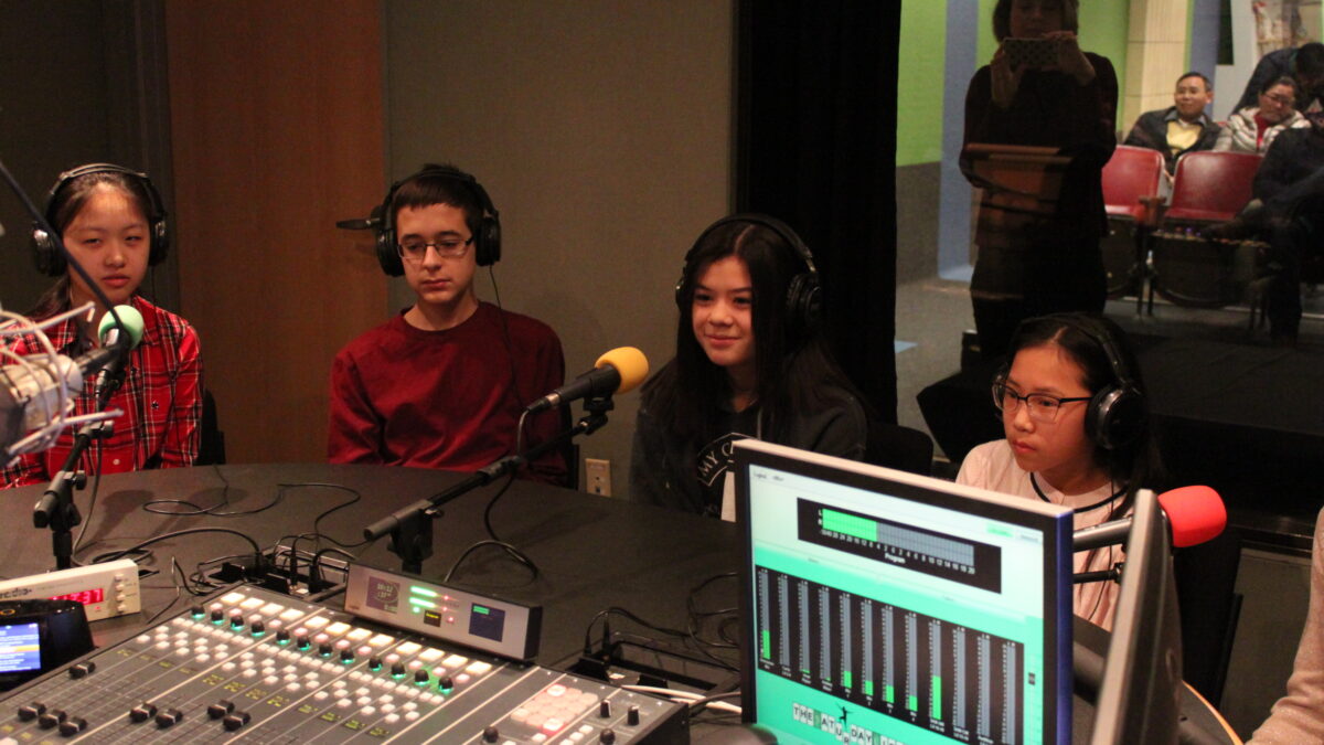 Four youth wearing headphones sitting behind microphones in a radio studio while an adult in the background takes a photo on their smartphone