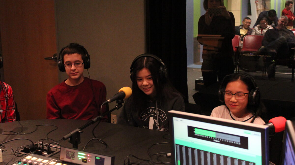 Three youth wearing headphones speaking into microphones in a radio studio while an adult in the background takes a photo on their smartphone