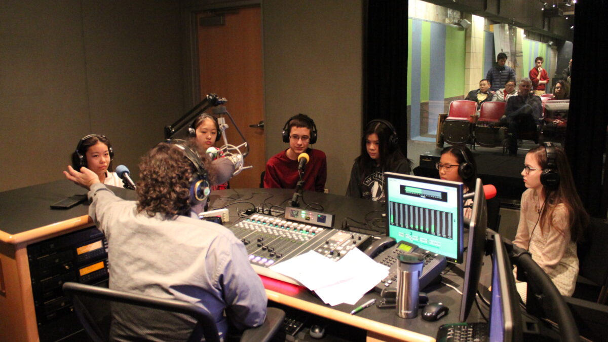 Six youth wearing headphones sitting behind microphones being interviewed by an adult wearing headphones sitting behind a microphone and a mixing board in a radio studio
