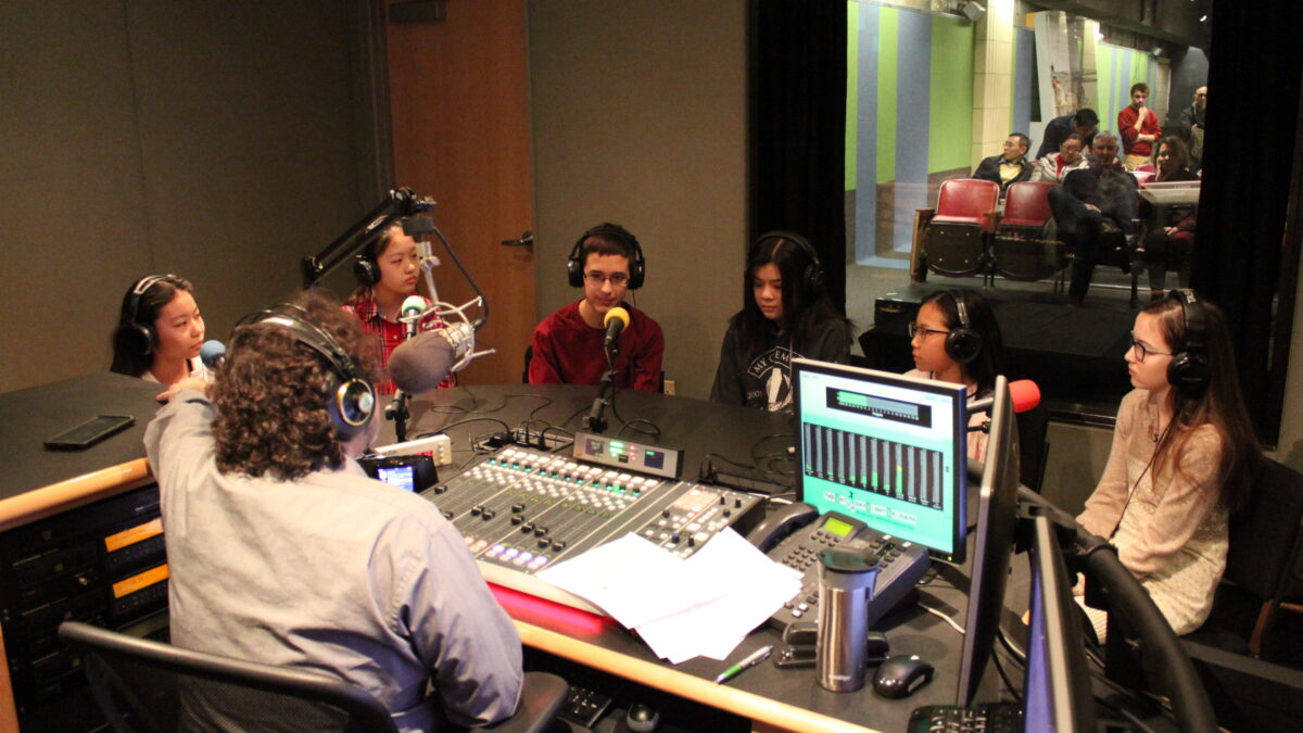 Six youth wearing headphones sitting behind microphones being interviewed by an adult wearing headphones sitting behind a microphone and a mixing board in a radio studio