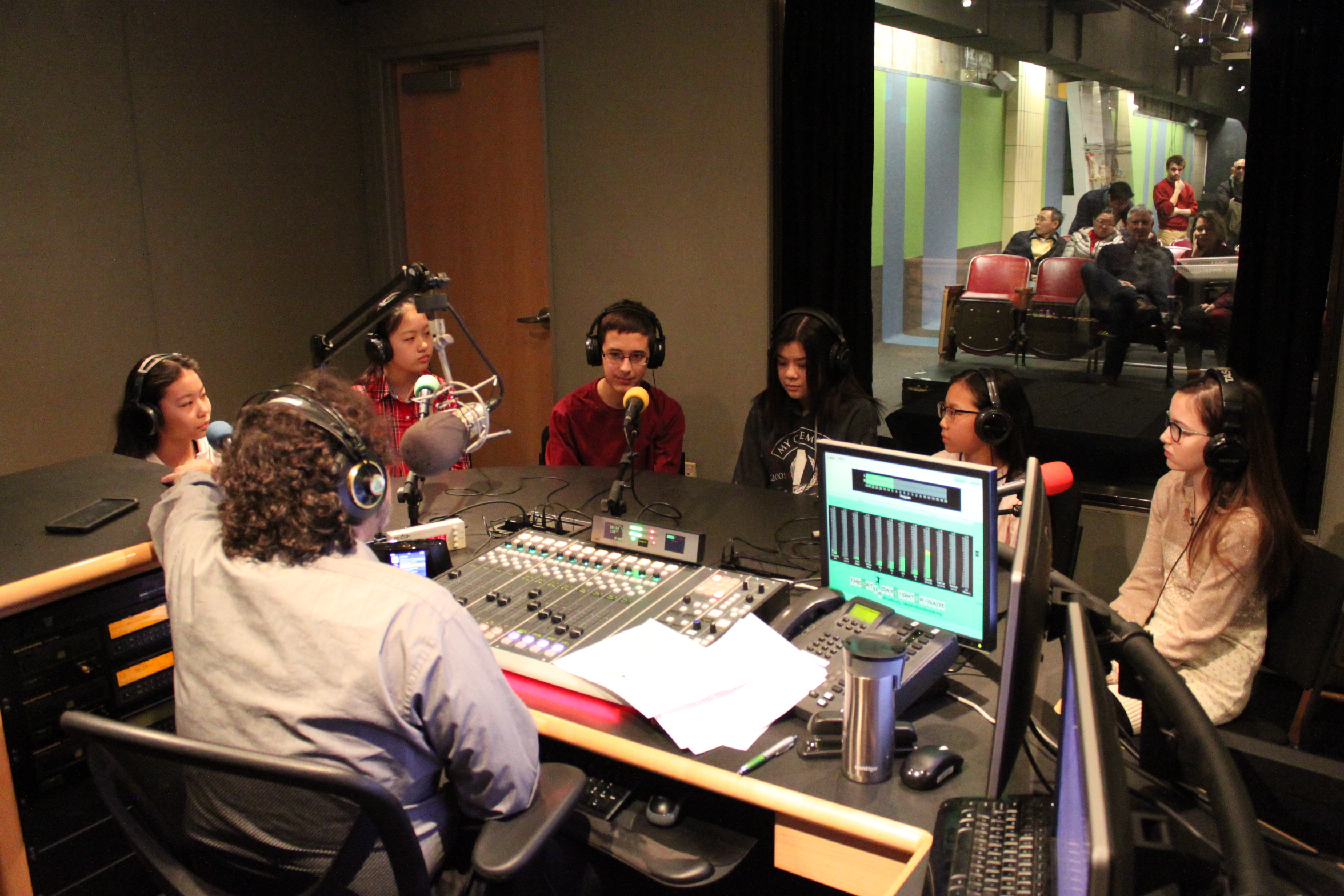Six youth wearing headphones sitting behind microphones being interviewed by an adult wearing headphones sitting behind a microphone and a mixing board in a radio studio