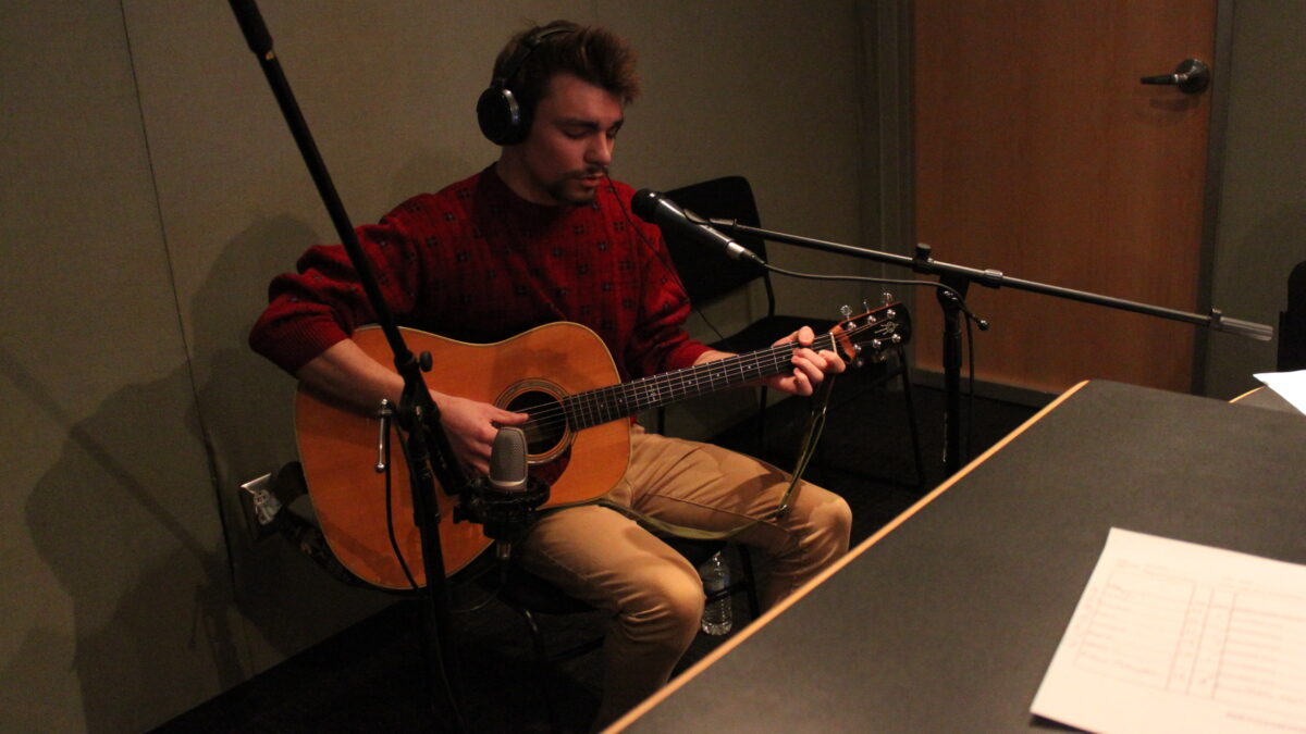 Individual wearing headphones playing guitar and singing into a microphone in a radio studio