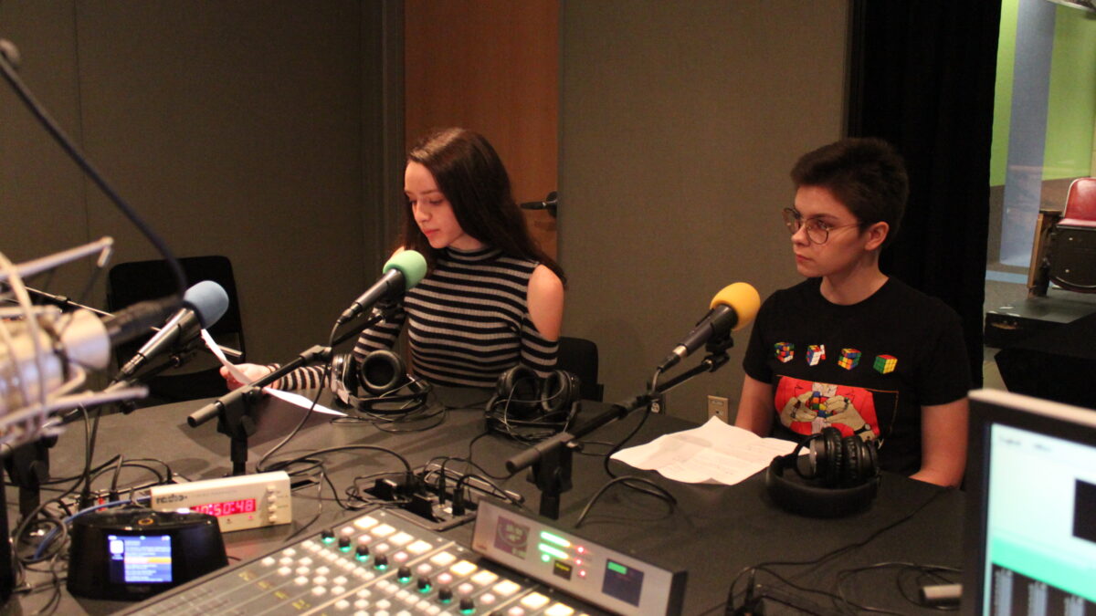 Two teens sitting behind microphones and looking at pieces of paper in a radio studio