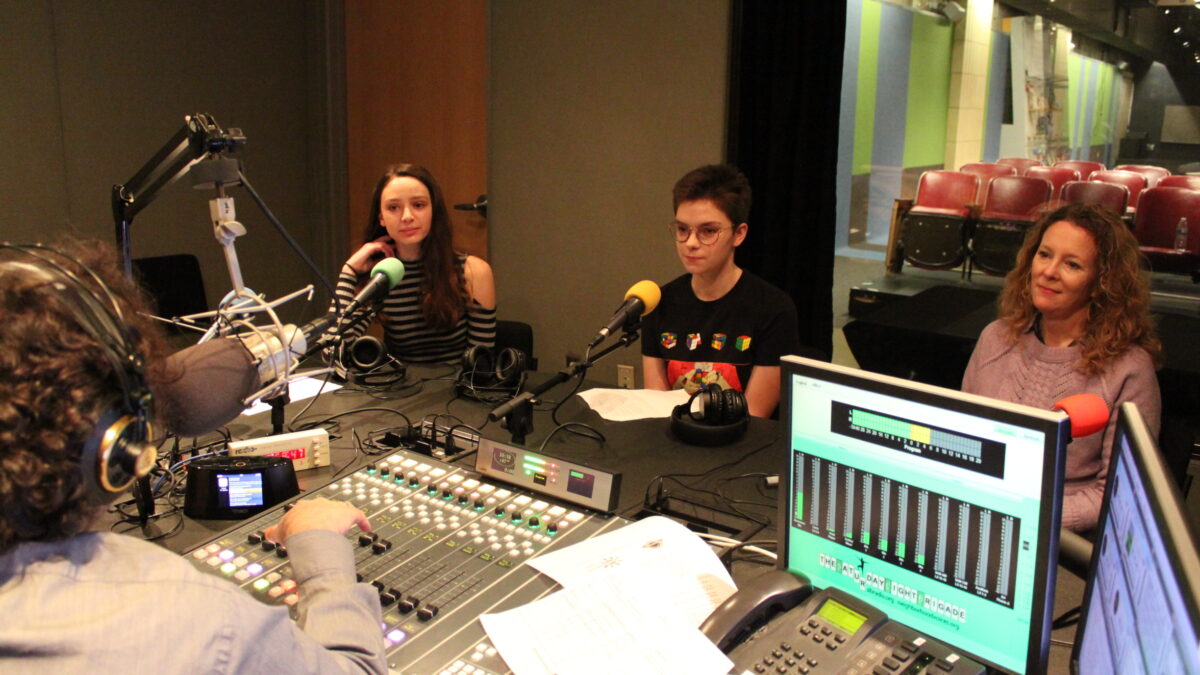 Two teens and an adult sitting behind microphones being interviewed by an adult wearing headphones speaking into a microphone sitting behind a mixing board in a radio studio