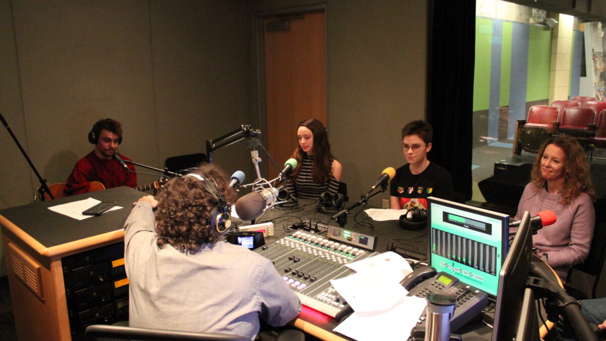 An individual wearing headphones and holding a guitar, two youth sitting behind microphones, and an adult sitting behind a microphone being interviewed by an adult wearing headphones speaking into a microphone sitting behind a mixing board in a radio studio