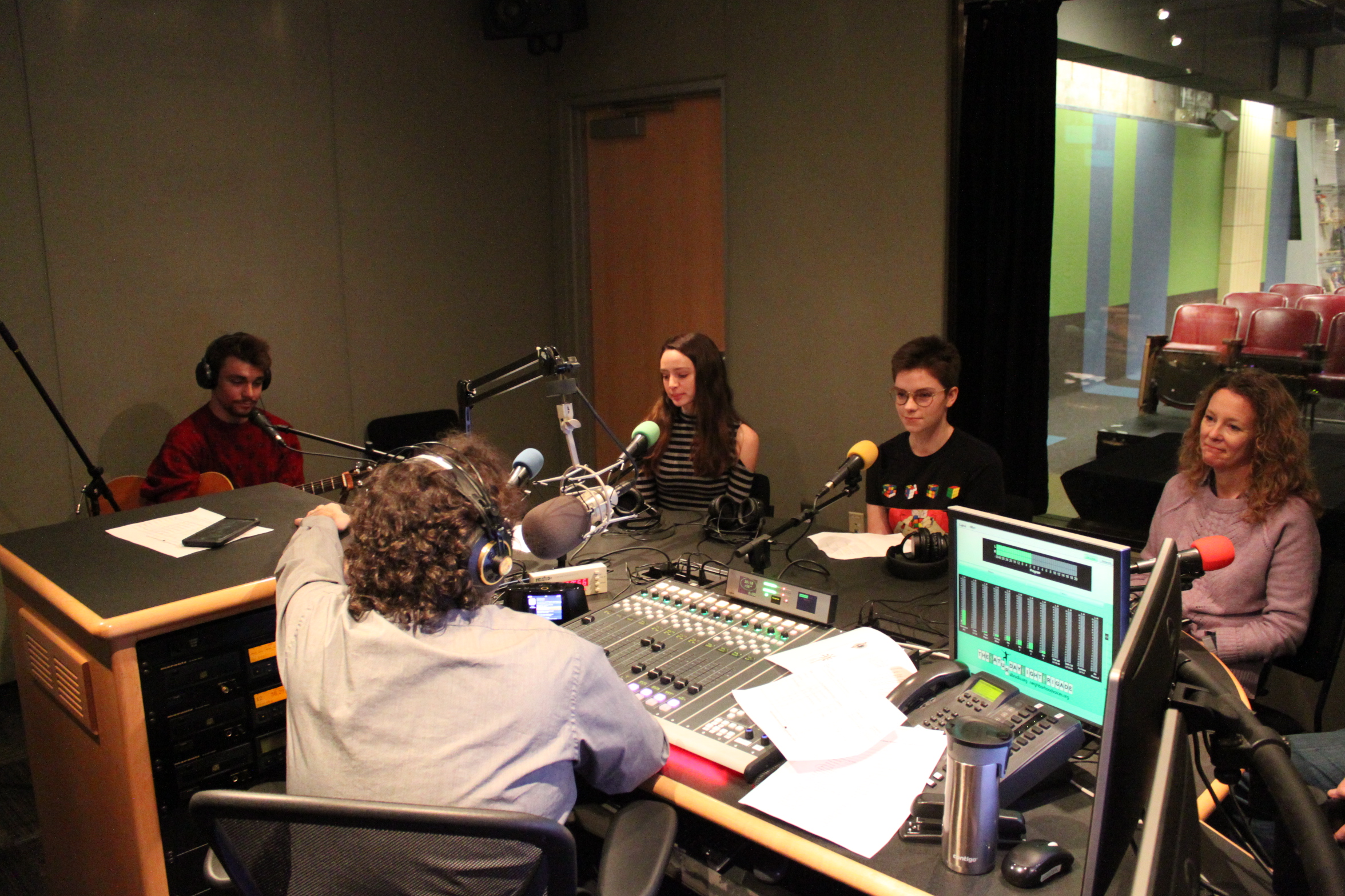 An individual wearing headphones and holding a guitar, two youth sitting behind microphones, and an adult sitting behind a microphone being interviewed by an adult wearing headphones speaking into a microphone sitting behind a mixing board in a radio studio