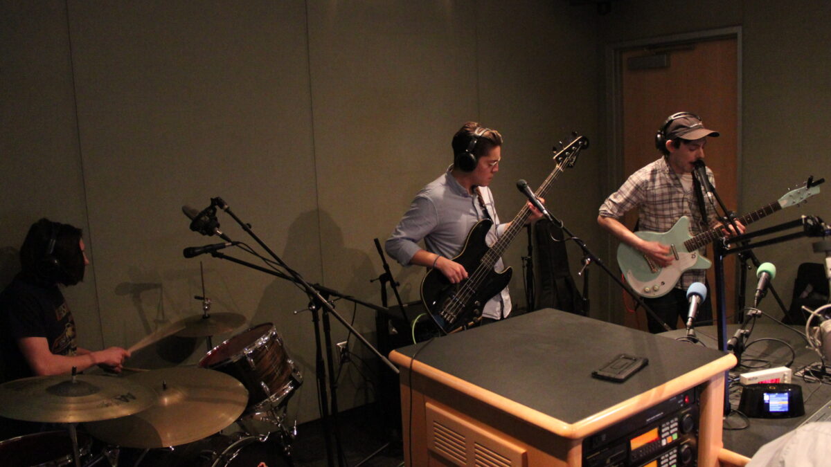 Three adults wearing headphones playing drum set, bass, and guitar in a radio studio, the guitarist singing into a microphone