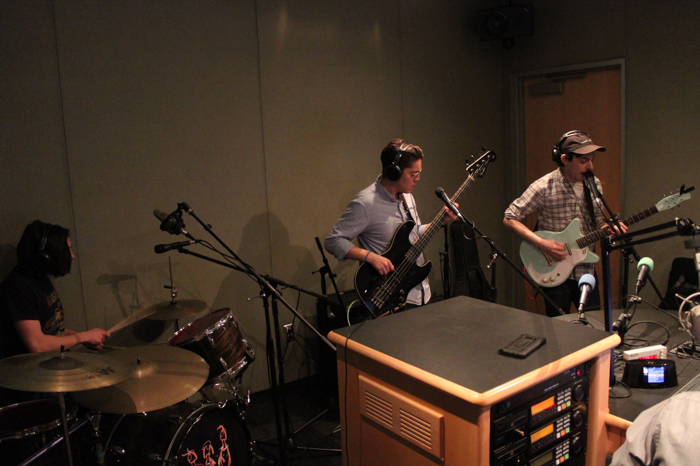 Three adults wearing headphones playing drum set, bass, and guitar in a radio studio, the guitarist singing into a microphone