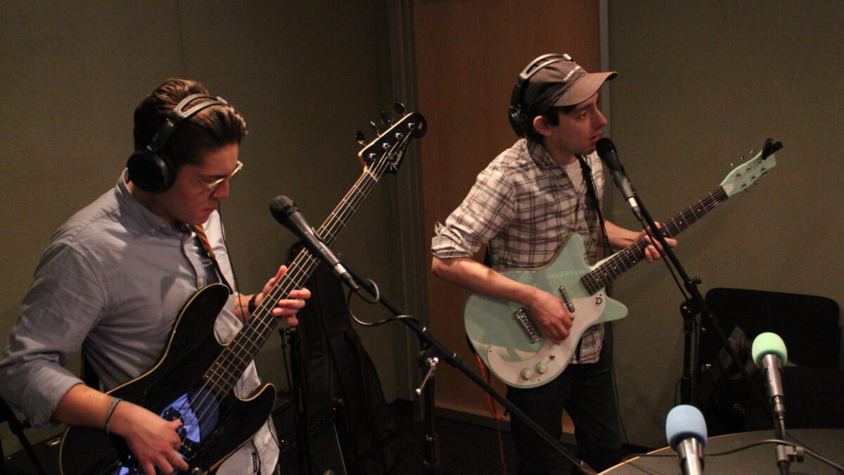 An adult wearing headphones playing bass and an adult wearing headphones playing guitar and singing into a microphone in a radio studio