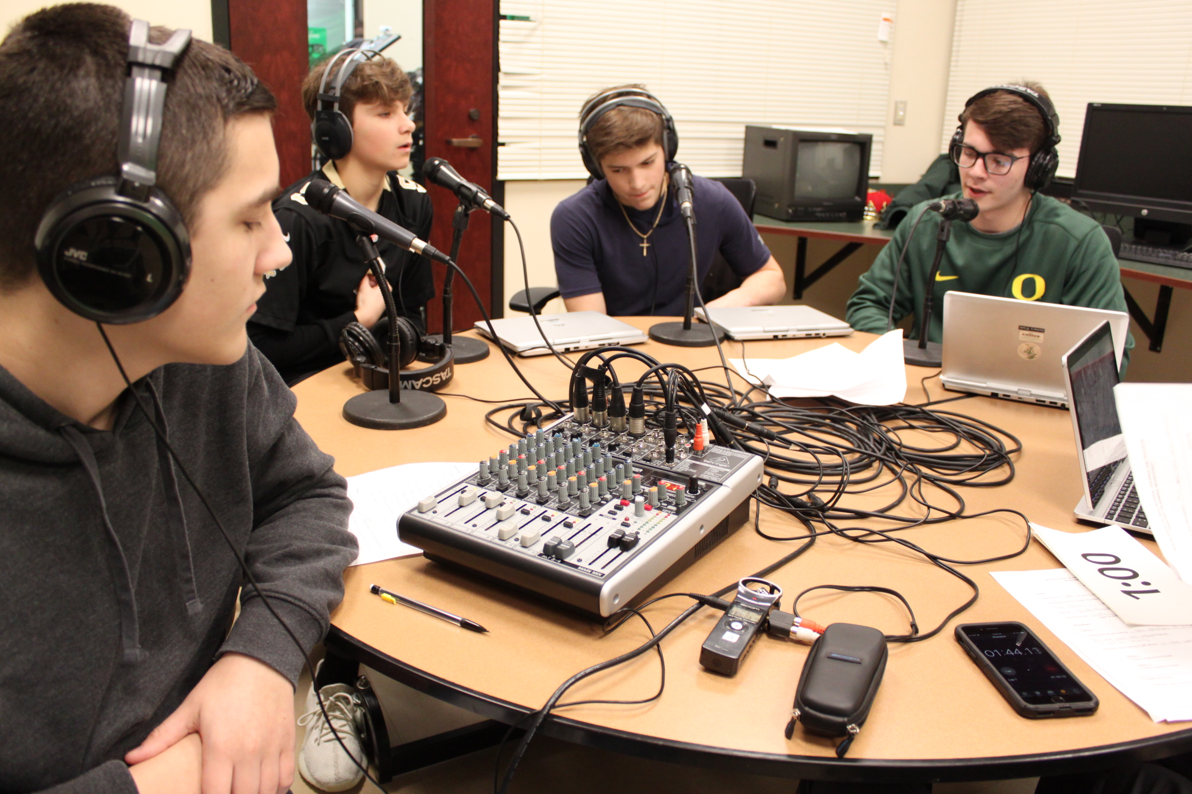 One teen wearing headphones listening to three teens wearing headphones speaking into microphones connected to a mixing board connected to a portable recorder