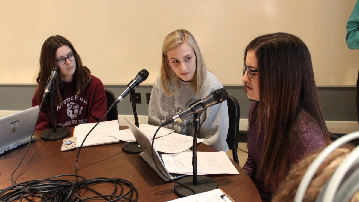 One teen speaking into a microphone while two teens sitting behind microphones look on