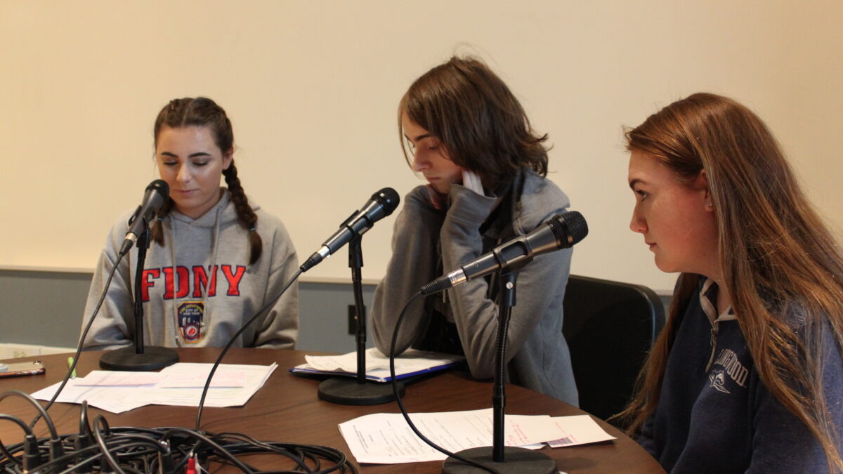 Three teens sitting behind microphones looking down at pieces of paper