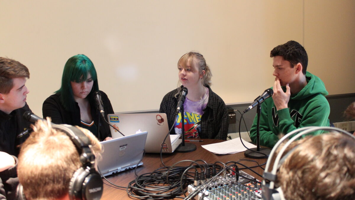 Two teens wearing headphones interviewing four teens speaking into microphones looking at two laptops