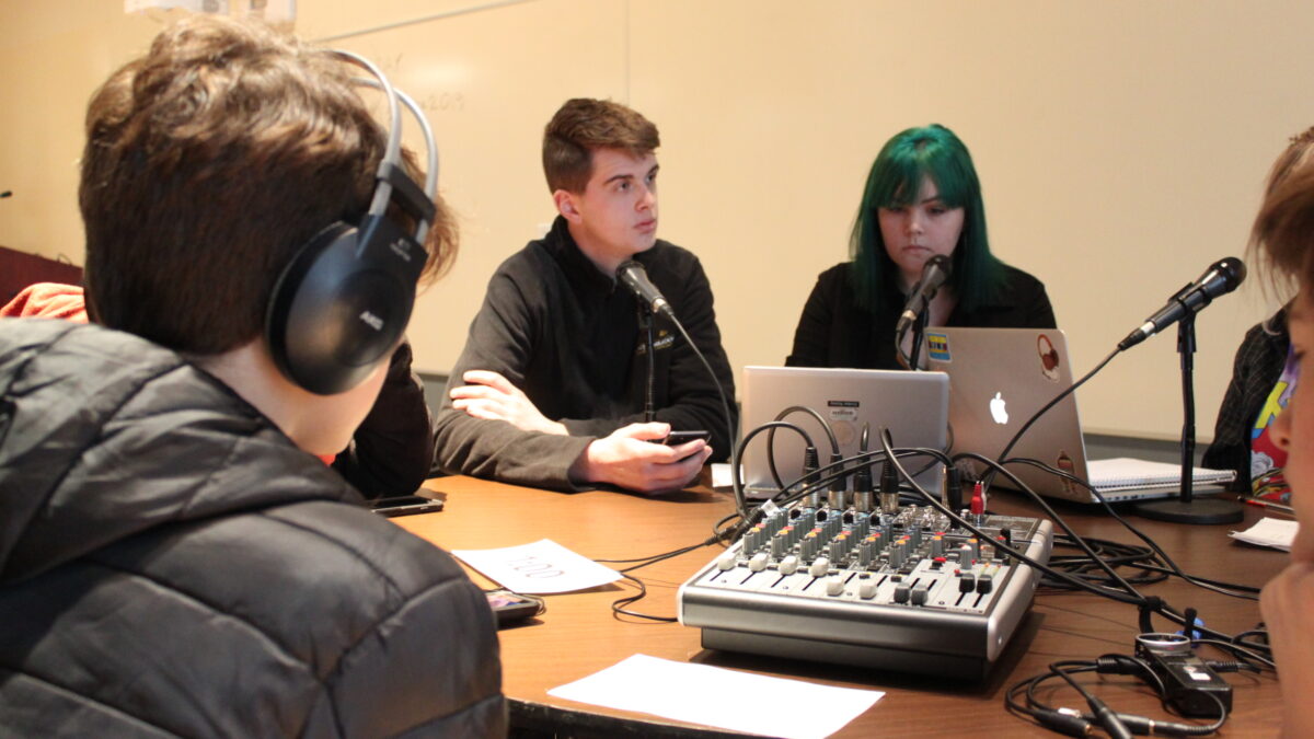 A teen wearing headphones interviewing three teens speaking into microphones and looking down at laptops and a smartphone