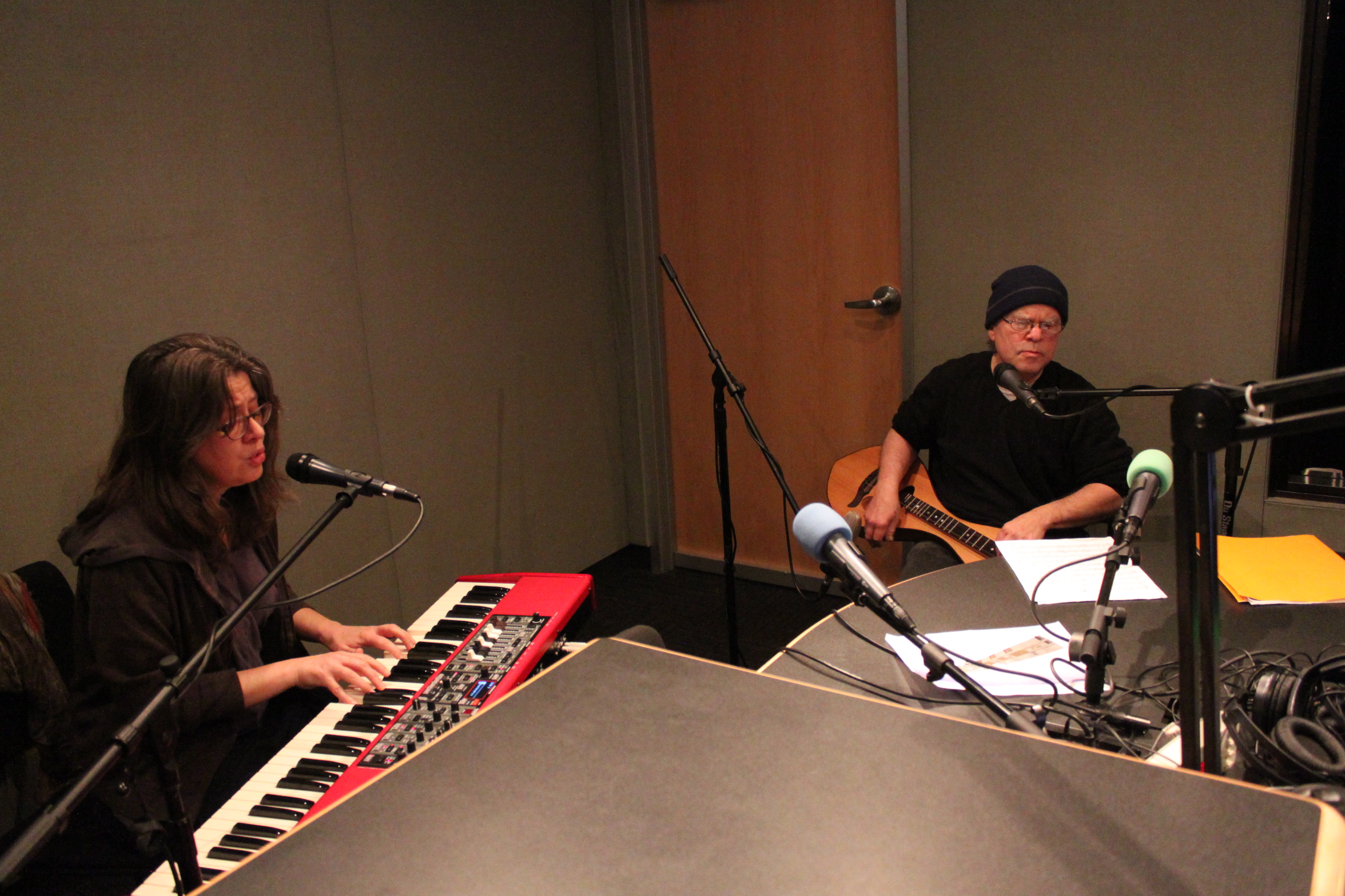 An adult playing a synthesizer and singing into a microphone, and an adult playing dulcimer in a radio studio