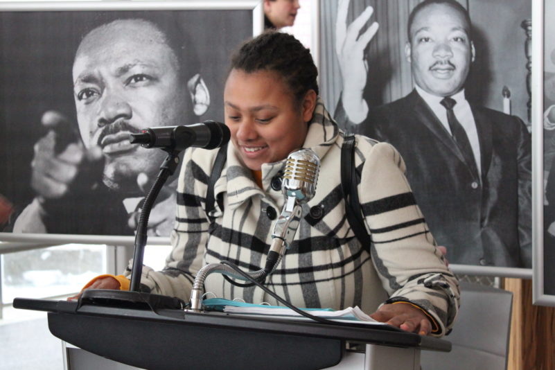 A teen speaking into two different styled microphones in front of two black and white images of an adult gesturing with their hands