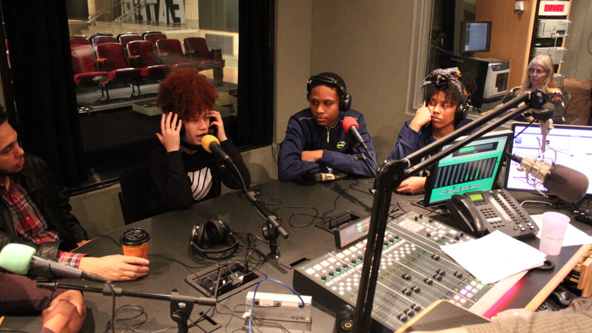 Four teens wearing headphones speaking into microphones in a radio studio
